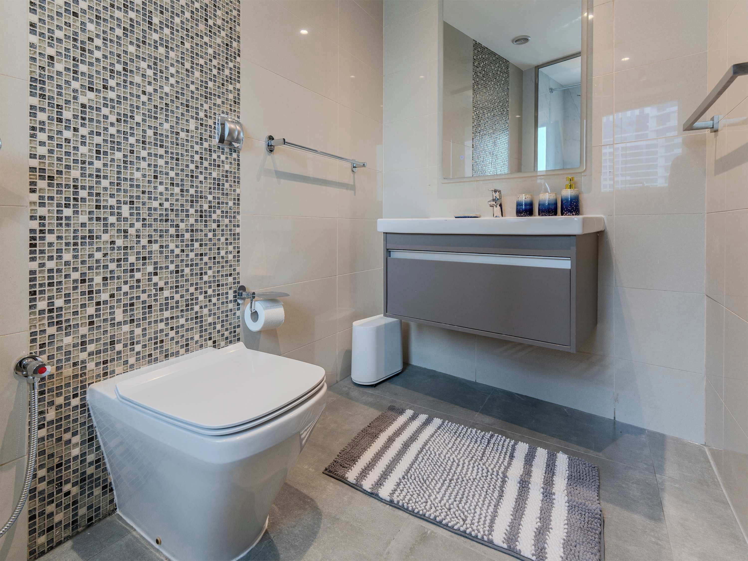 Une salle de bain moderne avec des toilettes, un lavabo surmonté d’un miroir et un mur recouvert de mosaïques.