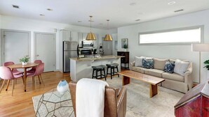 A modern kitchen with white cabinets, a stainless steel refrigerator, and a central island with stools. There is a beige sofa with patterned cushions in the living area, and a wooden coffee table.