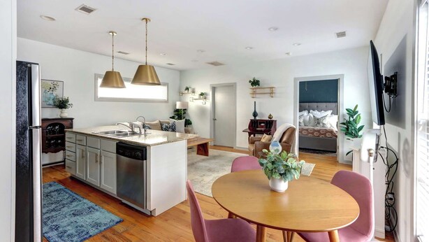 A modern kitchen with a central island, a dining area with a round table and chairs, and a bedroom with a bed and a TV.