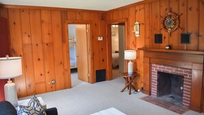 A wooden-paneled room with a fireplace, a bed in the background, and a lamp on a side table. - Light and bright two bedroom home in Harwich featuring central air! (West Harwich)