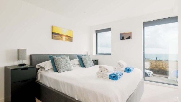 A modern bedroom with a large bed, a nightstand, a window with a view, and a painting on the wall. - Rise and Shine - With a Balcony On the Seafront (Christchurch)