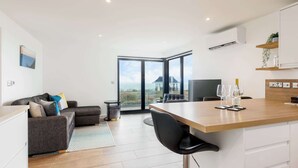 A modern living room with a sofa, a dining area with a table and chairs, and a large glass door leading to a balcony with a view of the sea. - Rise and Shine - With a Balcony On the Seafront (Christchurch)