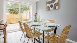 A dining area with a glass table and wicker chairs, a wooden fence outside, and a modern abstract painting on the wall. - Shell Cove - With Sunny Rear Patio Near Harbourside Park (Poole)