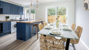 A modern kitchen with dark blue cabinets, a central island, and a dining area with wicker chairs and a glass table. - Shell Cove - With Sunny Rear Patio Near Harbourside Park (Poole)