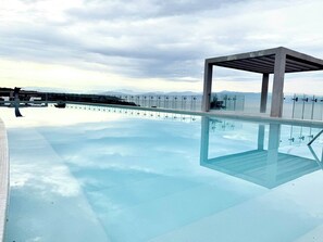 A clear infinity pool with a glass floor, a modern outdoor seating area, and a view of the sea and mountains in the background.