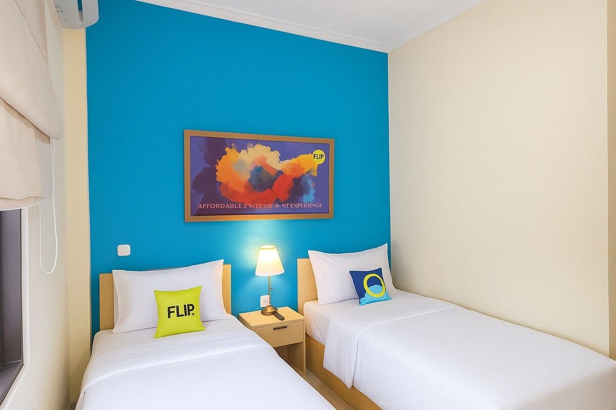 A hotel room with two beds, a nightstand with a lamp, a painting on the wall, and a door to the right.