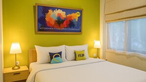 Habitación de hotel con cama, dos mesitas de noche con lámparas, un cuadro abstracto de colores en la pared y una ventana con persianas.
