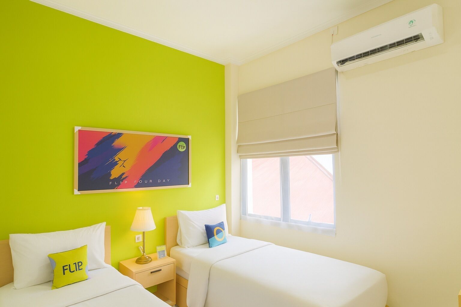 A room with two beds, a window with a beige blind, a wall-mounted air conditioner, and a colorful abstract painting.
