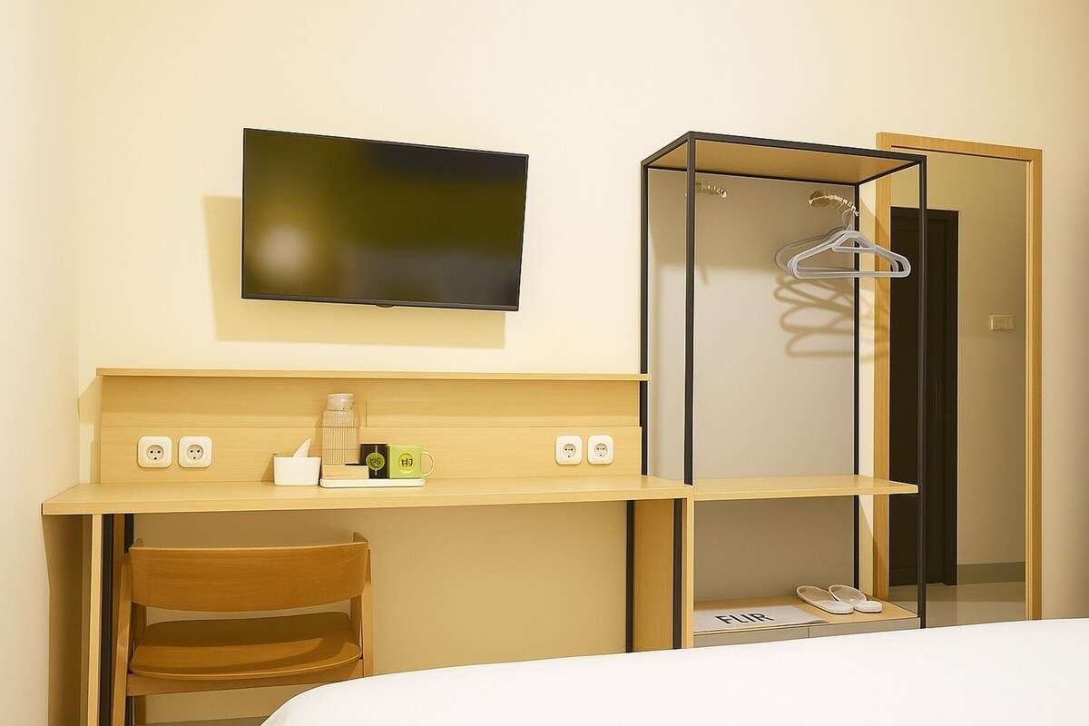 A hotel room with a bed, a desk with a chair, a TV mounted on the wall, a wardrobe with a mirror, and a hanging clothes rack.