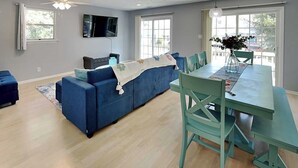 Un salon moderne avec un canapé bleu, une table à manger avec des chaises, un téléviseur à écran plat fixé au mur et de grandes fenêtres laissant entrer la lumière naturelle.