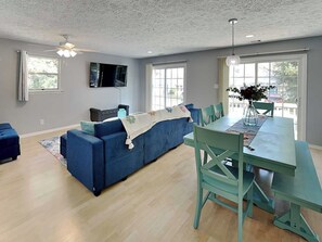 Un salon moderne avec un canapé bleu, une table à manger avec des chaises, un téléviseur à écran plat fixé au mur et de grandes fenêtres laissant entrer la lumière naturelle.