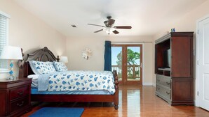 Une chambre avec un lit en bois, un ventilateur de plafond, une commode, un meuble pour la télévision et une vue sur l’extérieur grâce à une porte-fenêtre coulissante.