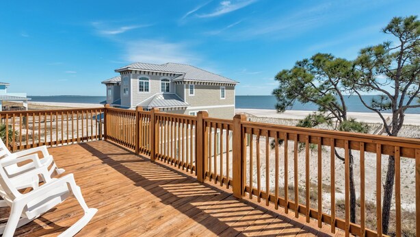 Une terrasse en bois avec des chaises blanches, une vue sur la plage et une maison avec un toit gris.