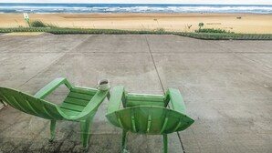 Twee groene stoelen met een kopje op de grond, een betonnen terras en een strand met golven op de achtergrond.