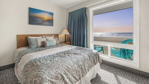 A hotel room with a bed, a painting of a sunset, a view of the beach, and a balcony with chairs. - Oceanfront Ground Floor Studio  - Pool & Hot Tub! (Lincoln City)