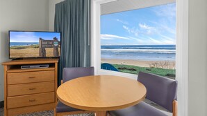 A hotel room with a television displaying a beach scene, a wooden table, two chairs, and a large window with a view of the ocean.