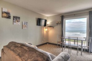 Een kamer met een groot raam dat uitzicht geeft op de oceaan, een flatscreen-tv aan de muur, een bank en een eettafel met stoelen.