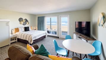 A hotel room with a bed, a TV, a dining area with chairs, and a view of the beach.