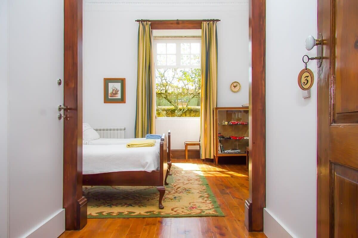 A bedroom with a bed, a window with curtains, a radiator, a clock, and a door with a number plate.