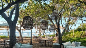 Outdoor seating with wicker furniture and cushions, surrounded by trees and umbrellas, overlooking a beach.