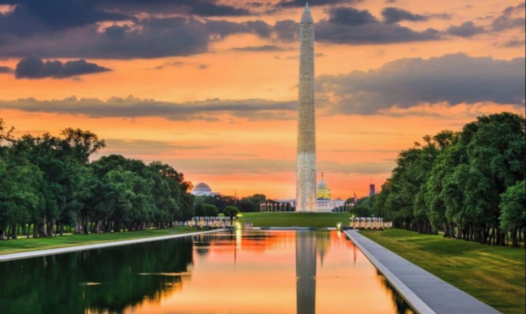 A tall monument with a reflection in a calm body of water, surrounded by trees and a clear sky.