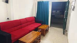 A living room with a red sofa, two wooden side tables, a black armchair, a teal curtain, and a visible door leading to another room. - Skyry Residency (Chennai)
