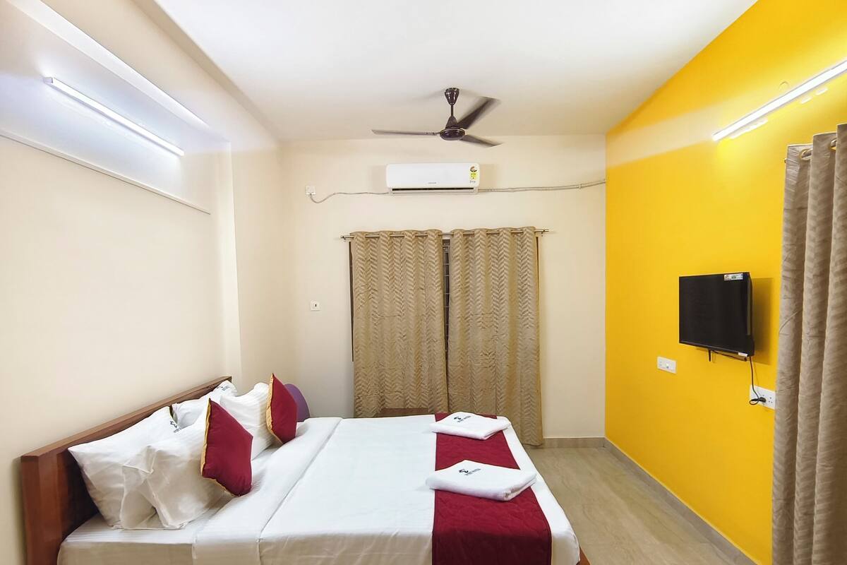 A neatly made bed with white and red bedding, a ceiling fan, an air conditioning unit, a television on the wall, and a window with curtains.