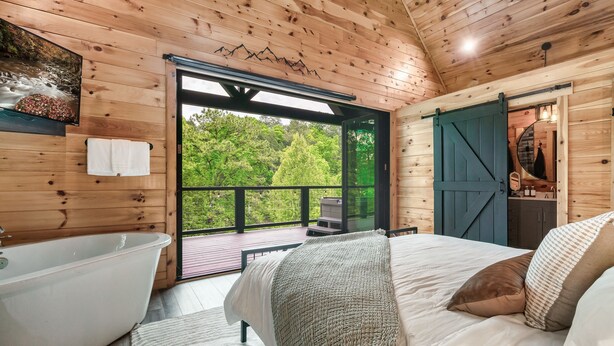 A wooden cabin bedroom with a large bed, a bathtub, and a view of greenery outside. - Romantic Couples Treehouse! Theatre! 3 min to PF! (Pigeon Forge)