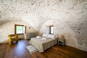 Une chambre avec un plafond en pierre, un plancher en bois, un lit avec du linge de lit blanc, une chaise en bois et une table de chevet avec une lampe.