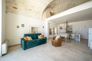 Un vaste espace de vie comprenant un canapé et un fauteuil, une cuisine à l’arrière-plan et un plafond haut avec des murs en briques apparentes.