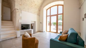 Una sala de estar con televisor, sofá y sillón. Se observa una escalera y un ventanal amplio.