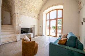 Un salon avec un téléviseur, un canapé et un fauteuil. On y trouve un escalier et une grande fenêtre.