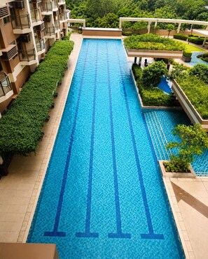 Una piscina larga con agua azul cristalina, rodeada de follaje y un edificio con balcones.