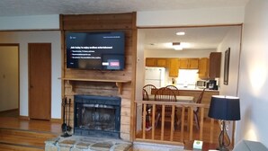 Ein Wohnzimmer mit Kamin, einem an der Wand befestigten Fernseher, einem Essbereich mit Holzsesseln und einer durch einen offenen Durchgang erkennbaren Küche.