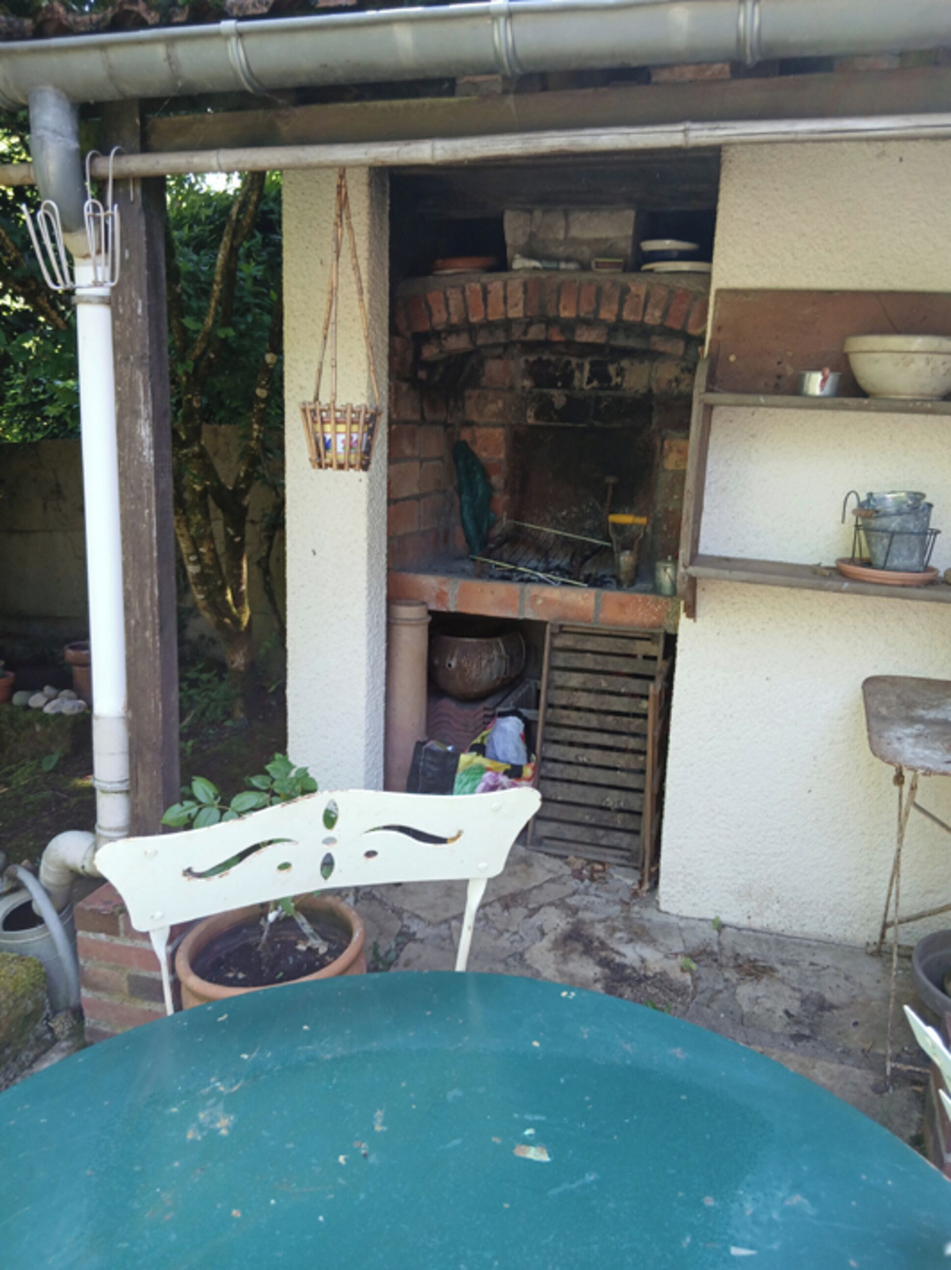 A rustic outdoor kitchen with a brick oven, a hanging basket, and a white metal chair.