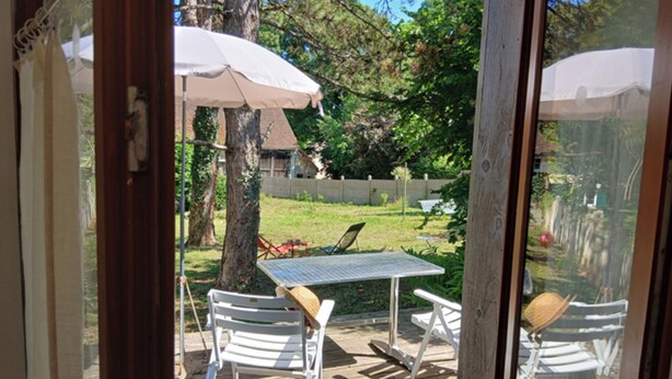 A wooden deck with a table and two chairs, an umbrella, and a view of a backyard with a lawn and trees. - Studio with terrace and garden 15 minutes from Guédelon (Treigny-Perreuse-Sainte-Colombe)