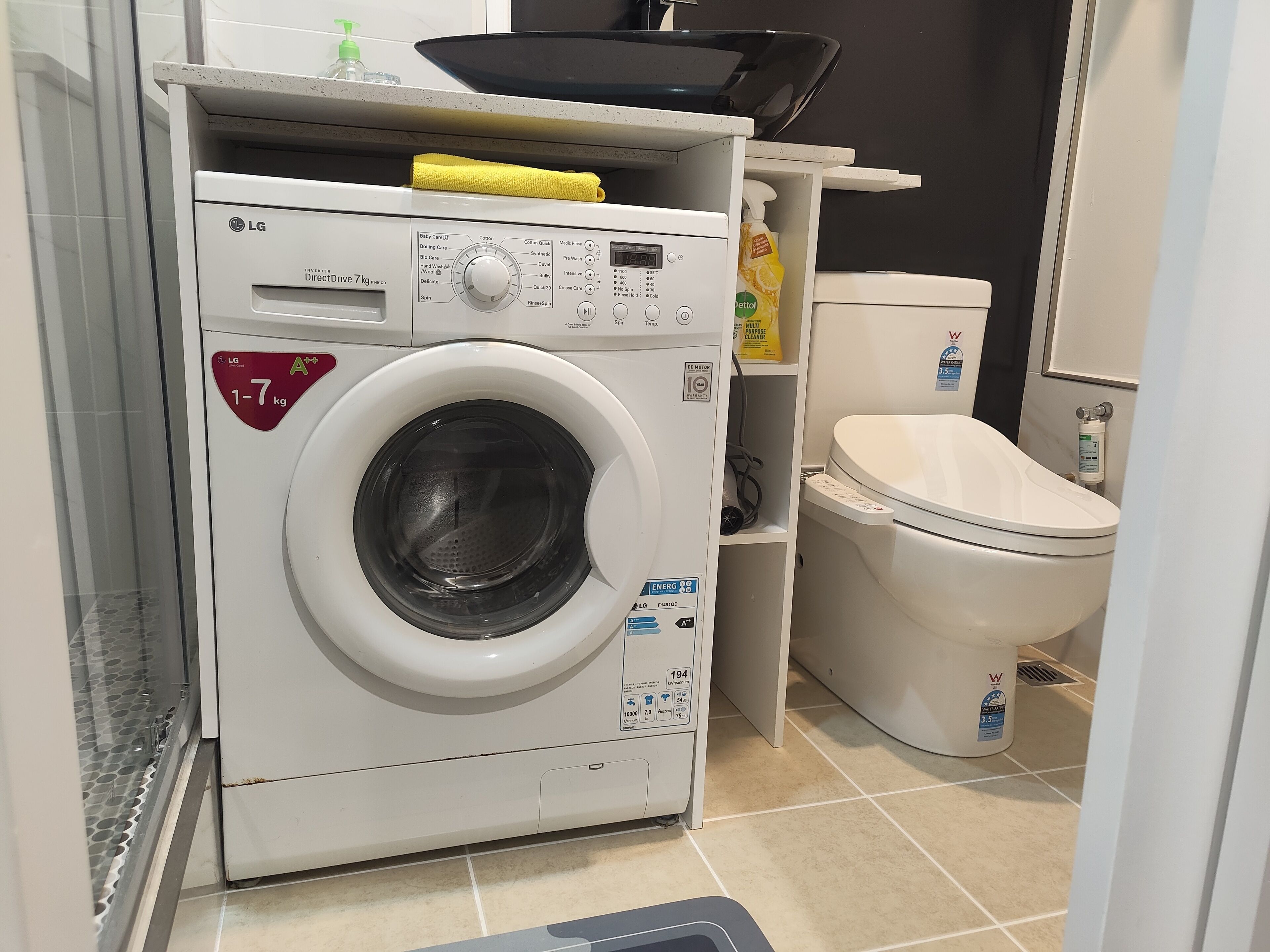 A compact laundry room with a front-loading washing machine, a toilet, and a shelf with cleaning supplies.