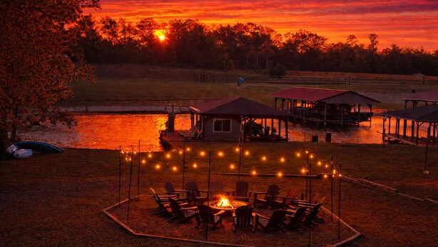 Zona de estar al aire libre junto al lago con sillas y una chimenea, rodeada de luces de cadena.