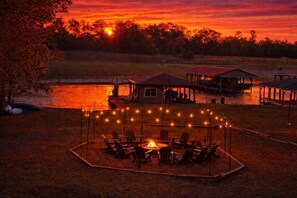 A lakeside outdoor seating area with chairs and a fire pit, surrounded by string lights.