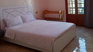 Un dormitorio con cama, cuna, ventana con vista a edificios y una lámpara en el techo.