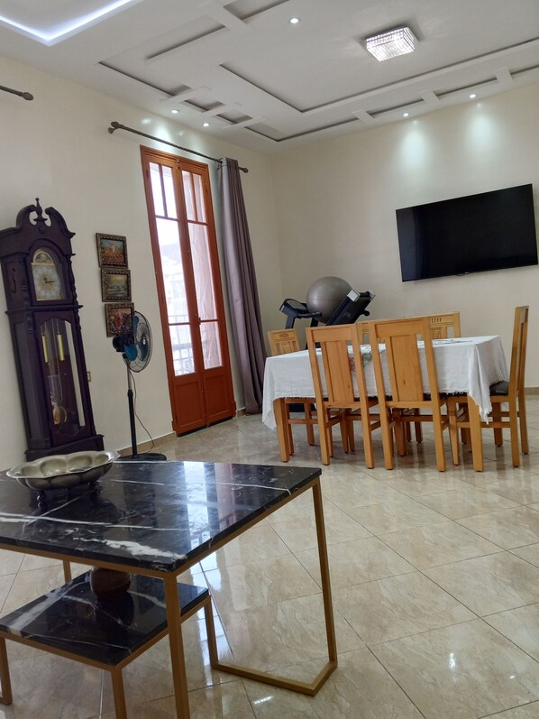A dining area with a marble table, wooden chairs, a ceiling fan, a clock, and a TV on the wall.
