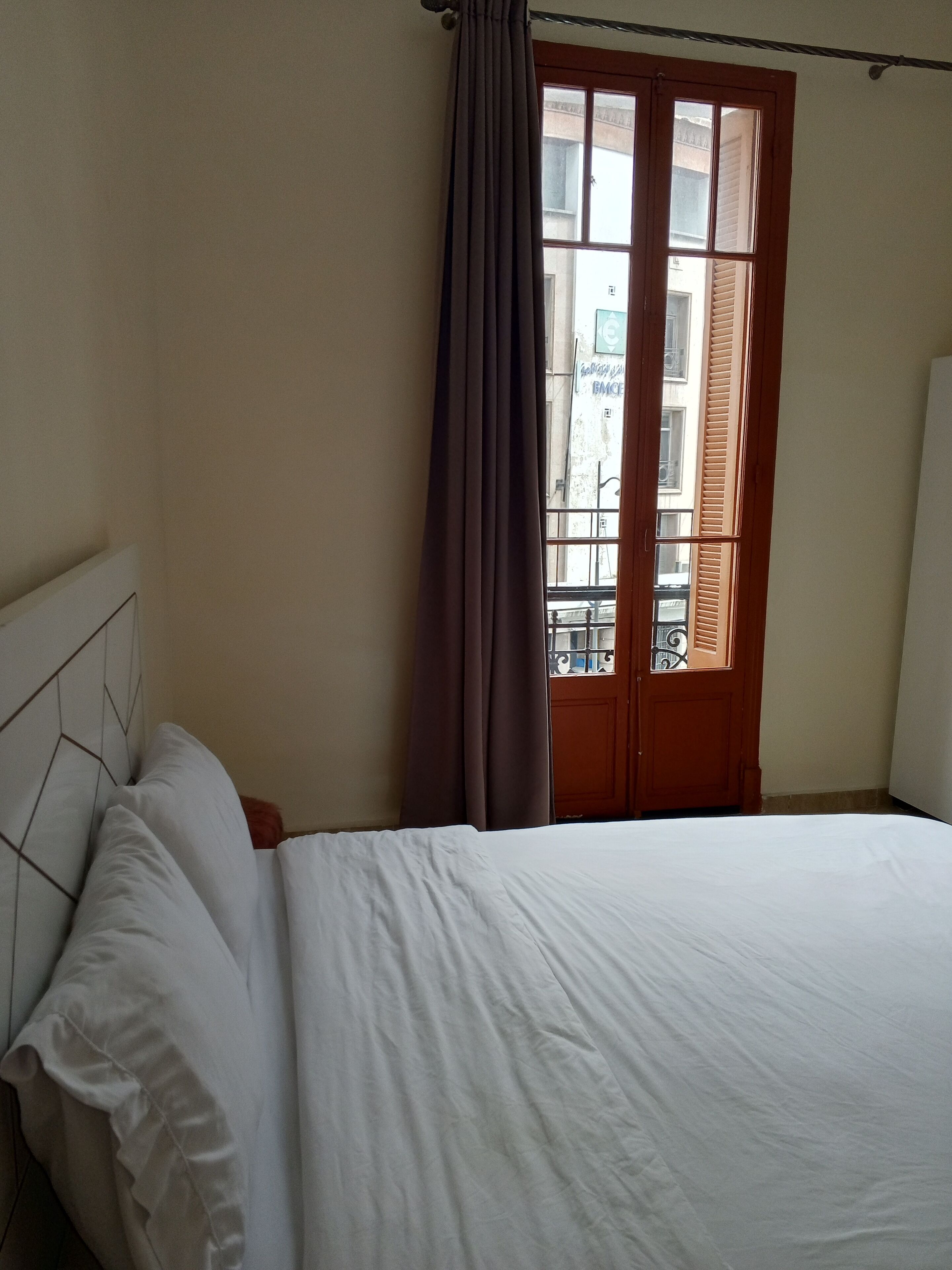A bedroom with a bed, a window with brown shutters, and a view of buildings outside.