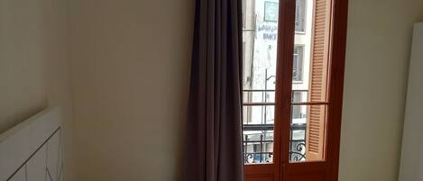 A bedroom with a bed, a window with brown shutters, and a view of buildings outside.