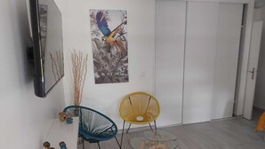 Una sala de estar moderna con un televisor de pantalla plana, un cuadro de un loro, dos sillas, una mesa de centro de vidrio y un estante pequeño con adornos.