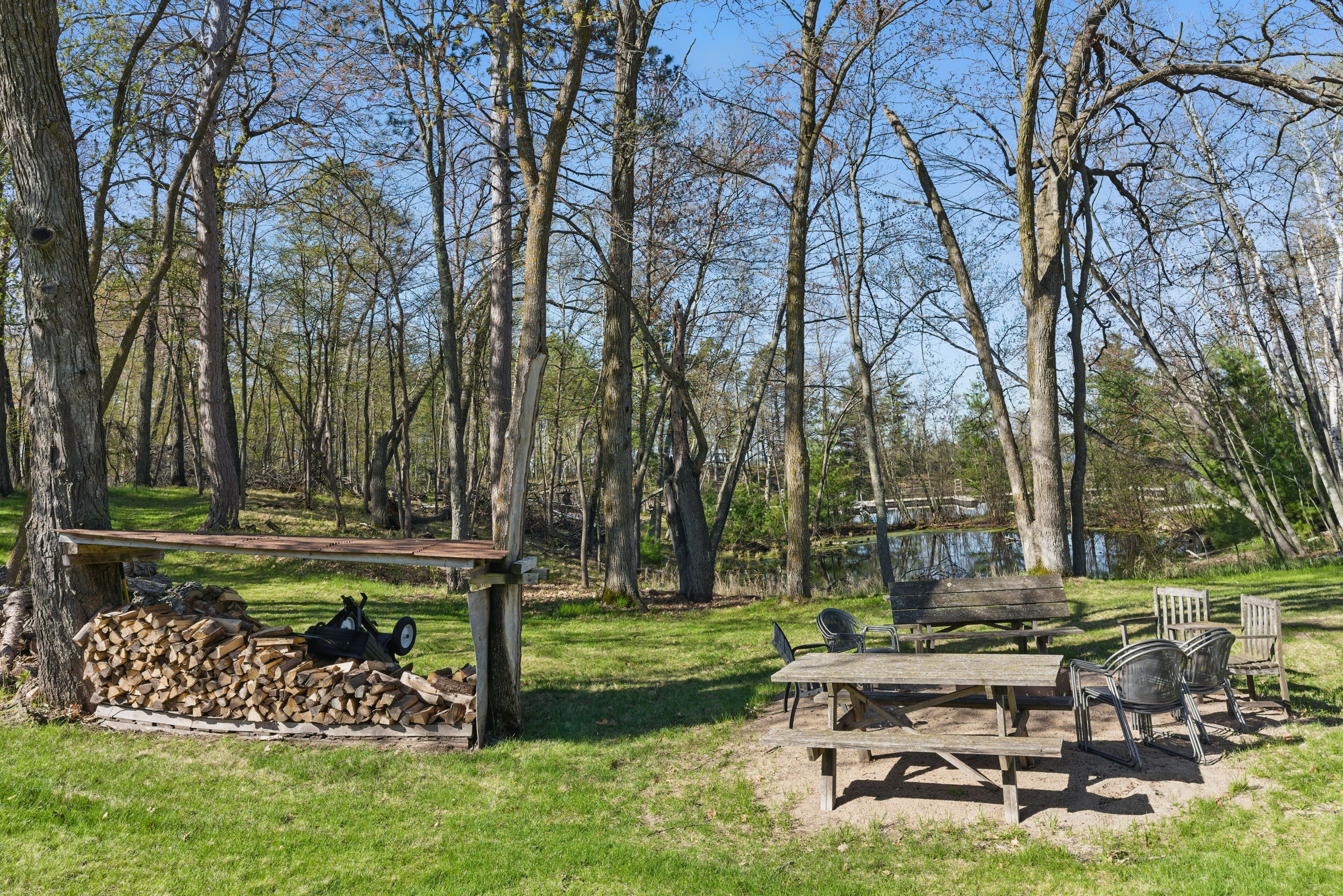 A picnic area with benches and chairs, surrounded by trees and a lake.