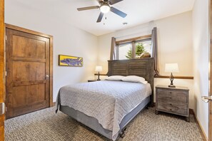 Ein Schlafzimmer mit Bett, Nachttisch, Holz-Kopfteil, Fenster mit Vorhängen, Deckenventilator und einer Tür.