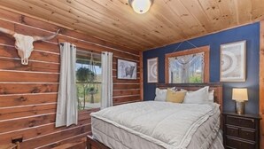 A wooden cabin bedroom with a bed, curtains, a nightstand, and a cow skull decoration.