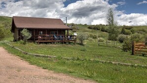 A rustic log cabin with a porch and outdoor seating, surrounded by a grassy landscape and a dirt path.