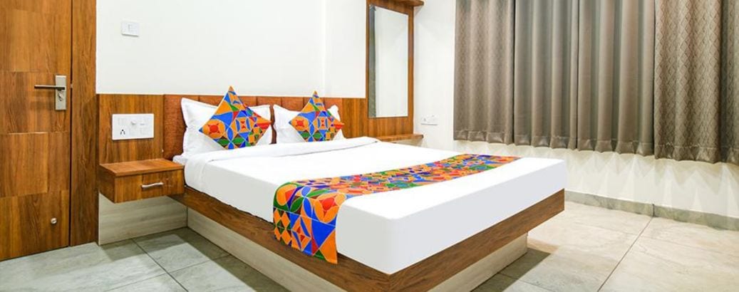A modern hotel room with a wooden headboard, a large bed with colorful bedding, a bedside table, and a window with curtains.