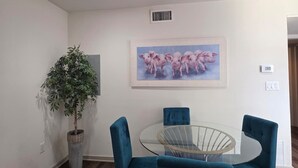 Un coin repas avec une table en verre entourée de quatre chaises de couleur turquoise, une plante en pot et un tableau représentant des cochons, accroché au mur.